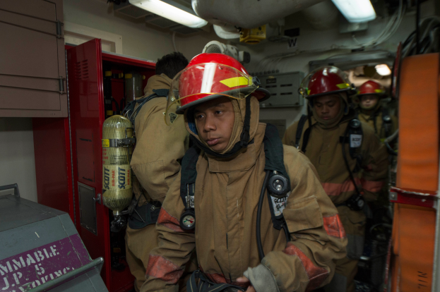 Eine Gruppe von Feuerwehrleuten in Uniformen und Helmen, einer hält einen Feuerlöscher, mit einem metallischen Objekt links und Lampen, Rohren und anderen Gegenständen im Hintergrund.