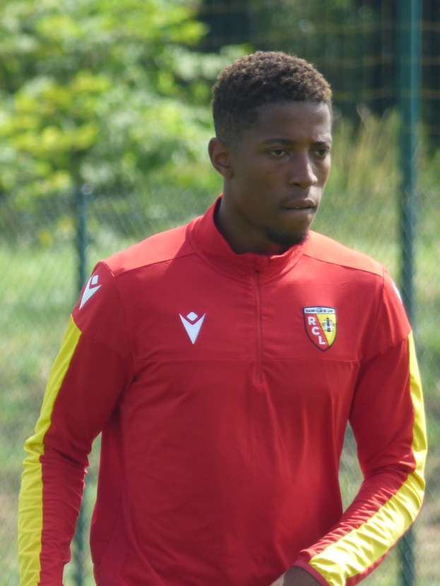 Ein junger Mann in einem roten und gelben T-Shirt steht auf einem Fußballfeld umgeben von einem Zaun und Bäumen und sieht aus wie ein Flügelstürmer mit entschlossener Miene.