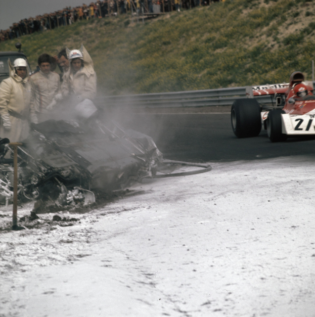 Ein verunglücktes Auto am Rand einer Rennstrecke, umgeben von einem Zaun und einer Gruppe von Menschen, mit Schnee auf dem Boden und einem Hügel und einem klaren blauen Himmel im Hintergrund.