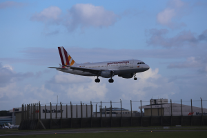 Germanwings Airbus A320-200 beim Abheben vom Frankfurt Airport mit Gras darunter, einem Zaun im Vordergrund, Gebäuden und Bäumen im Hintergrund und einer bewölkten Himmel darüber.