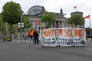 Eine Gruppe von Menschen marschiert eine von Bäumen gesäumte Straße entlang und hält ein Banner mit der Aufschrift 'Kampf für Solidarität', mit parkenden Fahrzeugen und einem mehrstöckigen Gebäude im Hintergrund unter einem bewölkten Himmel.